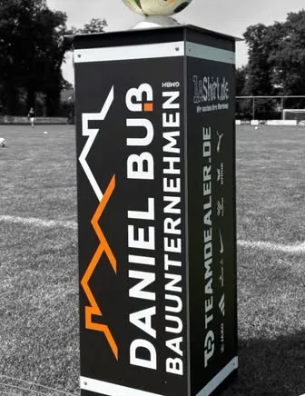 Werbepylon Daniel Buß Bauunternehmen am Rand eines Fußballplatzes, mit Logos von 1aShirt und Teamdealer
